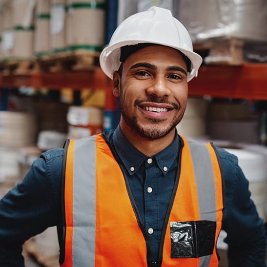 A man in a hardhat and safety vest over a button-down shirt stands in a busy warehouse. 