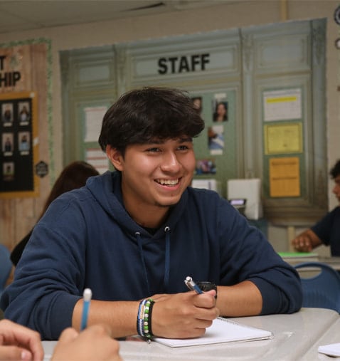 A smiling young person sitting at a table with a pen.