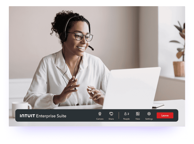 A woman in a headset sits at a desk and has a conversation on her laptop. 